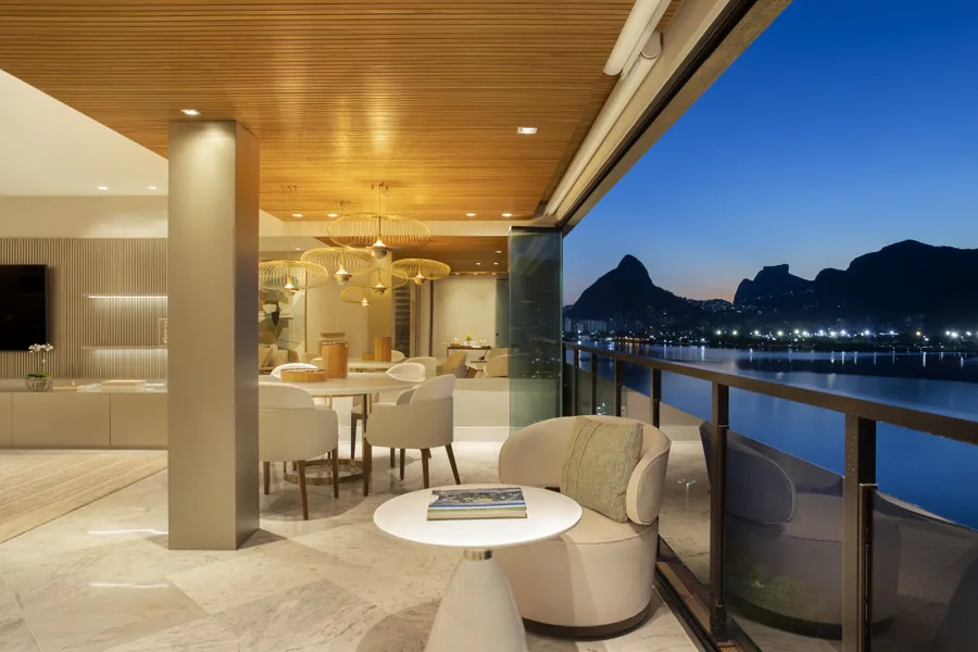 Varanda integrada à sala, com forro de madeira ripada e vista para a Lagoa ao anoitecer