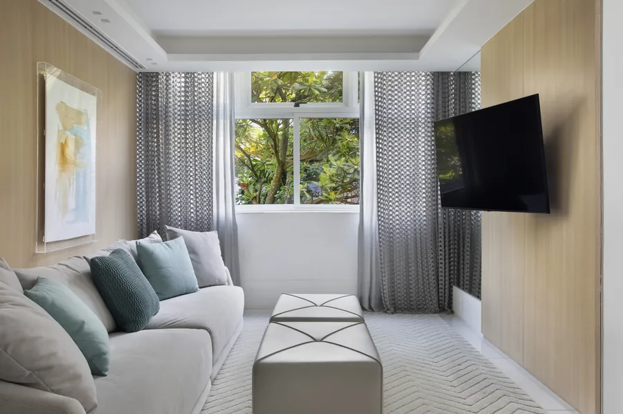 Sala de TV com painel em madeira clara, sofá e janela com cortinas
